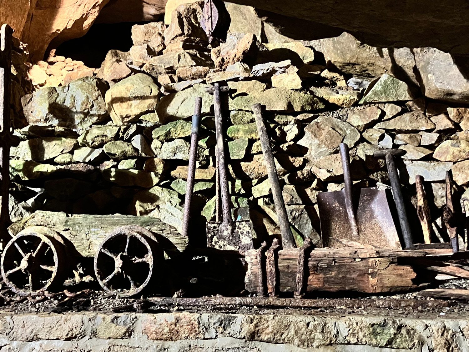 Old mine carts and tools beside a stone wall.